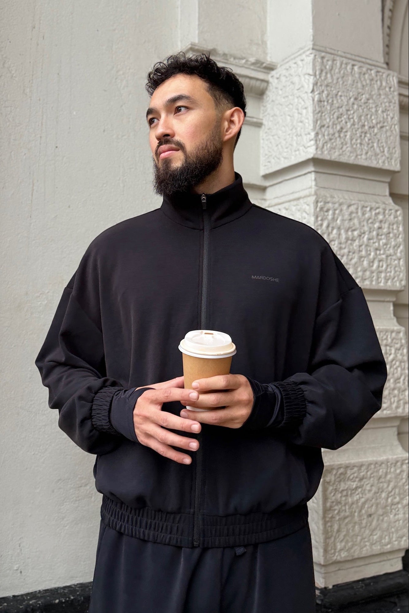 Man in black outfit holding a coffee cup against a light-colored building.
